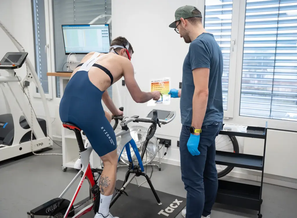 Sportler mit Atemmaske auf dem Ergometer, ein Mitarbeiter kontrolliert die Messung